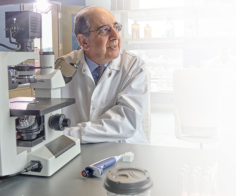Dr. Jose-Alain Sahel in a lab coat sits at a table with a microscope and lab equipment in a laboratory setting.