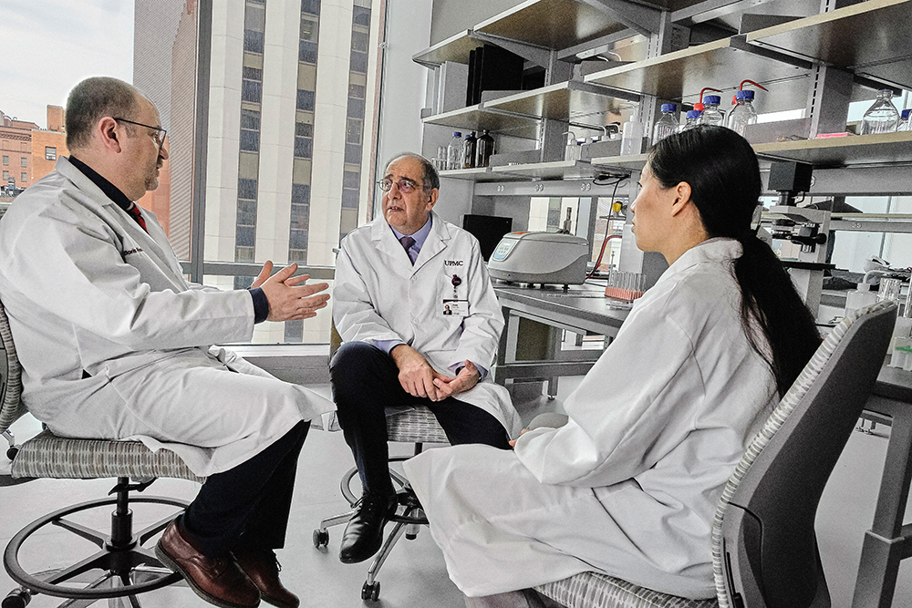 Dr. Jose-Alain Sahel and two people  in lab coats are seated and engaged in a discussion in a laboratory setting. Shelves with various lab equipment are visible in the background.