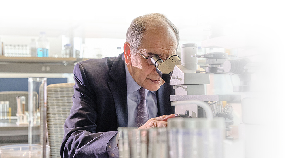 Dr. Jose-Alain Sahel dressed in a suit is using a microscope in a laboratory setting. Various laboratory equipment, such as glass beakers, are visible in the foreground, with shelves and bottles in the background.